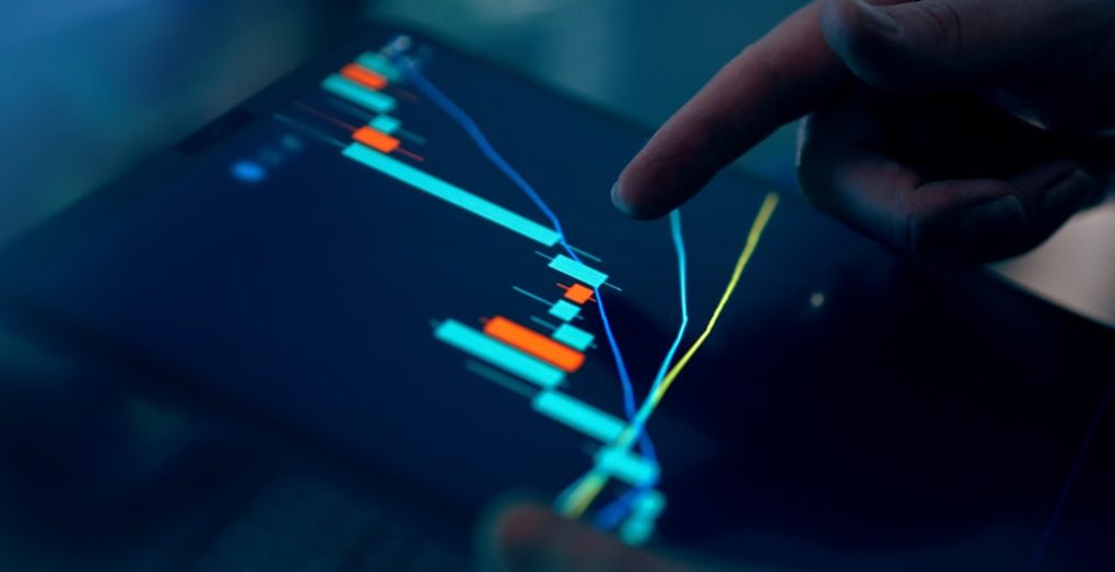 Close-up of a finger pointing at a digital trading chart with colorful candlestick patterns on a tablet screen.