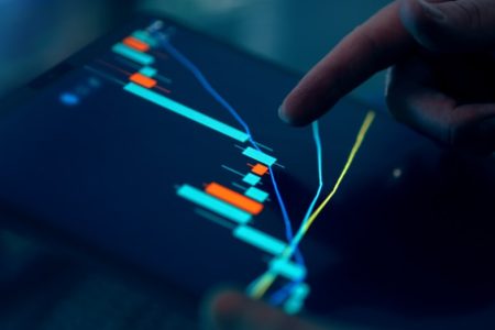 Close-up of a finger pointing at a digital trading chart with colorful candlestick patterns on a tablet screen.